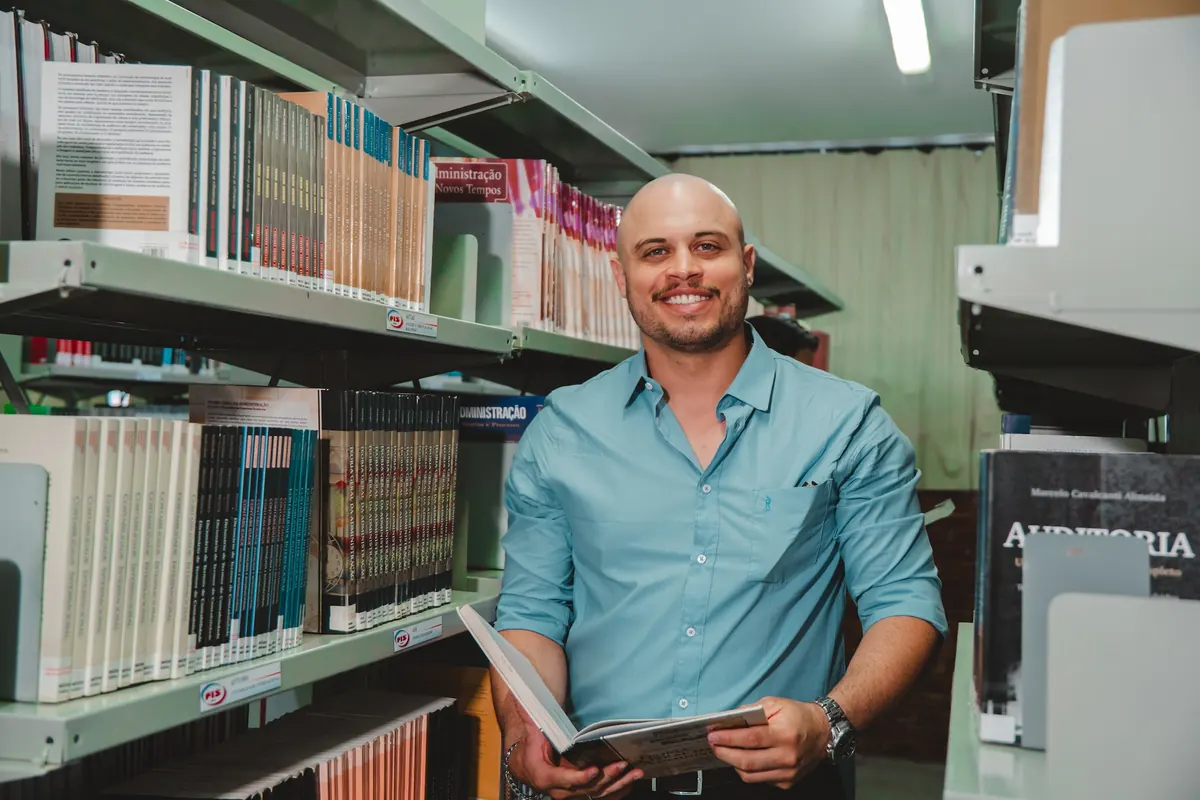 Homem de camisa azul sorrindo entre as estantes da biblioteca da UNIFIS, com um livro aberto nas mãos